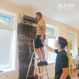 Alkar Professional - Dwóch monterów instaluje klimatyzator ścienny w pomieszczeniu z oknami, używając drabiny. Jeden z nich trzyma przewód. Widoczne logo firmy Alkar Professional.