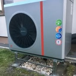 Alkar Professional - Szara pompa ciepła z czerwonym paskiem, zamontowana na metalowej konstrukcji z kamieniami, widoczna z boku na tle ściany budynku.