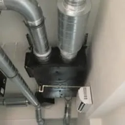 Widok instalacji wentylacyjnej z kanałami, centralą i klimatyzatorem, zamontowanej pod sufitem, z widocznymi elementami mocującymi i przewodami.