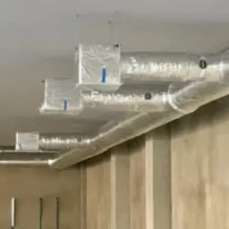 Szczegółowe ujęcie instalacji wentylacyjnej z aluminiowymi rurami i białymi skrzynkami na suficie, w surowym wnętrzu z beżowymi ścianami. Widok z dołu.