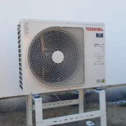 Biała pompa ciepła Toshiba na metalowym stelażu przed ścianą budynku. Widoczna tabliczka znamionowa i logo UPRO. Zewnętrzna jednostka klimatyzacji.