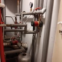 Złożona instalacja hydrauliczna z rurami, zaworami, manometrem i pompą obiegową, umieszczona przy ścianie. Widoczne elementy miedziane i stalowe, oświetlenie górne.