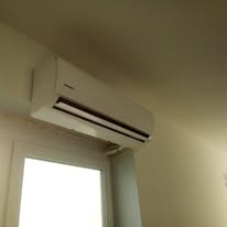 Biały klimatyzator ścienny zamontowany nad oknem. Widok od dołu, na tle białej ściany i sufitu. Urządzenie o prostym, nowoczesnym designie.