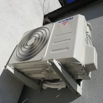 Biała jednostka zewnętrzna klimatyzacji Kaisai zamontowana na szarej ścianie budynku, widoczna z dołu pod kątem, z widocznymi rurami i wspornikami.