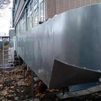 Szara, metalowa instalacja wentylacyjna na zewnątrz budynku, widoczna drabina i fragment zieleni. Ujęcie z dołu, ukazujące konstrukcję wsporczą instalacji.