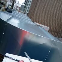 Długi, metalowy kanał wentylacyjny biegnący wzdłuż ściany budynku z cegły. Widoczne elementy montażowe i biała jednostka klimatyzacyjna na ścianie.