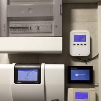 Panel sterowania instalacją grzewczą z wyświetlaczami LCD, zamontowany na ścianie obok skrzynki elektrycznej. Widoczne elementy instalacji i okablowanie.