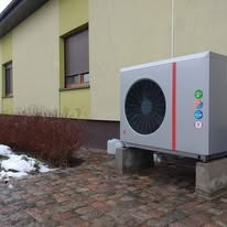 Szara pompa ciepła zamontowana na betonowych blokach przy ścianie budynku z brązowymi roletami i resztkami śniegu. Widok z boku, lekko z dołu.