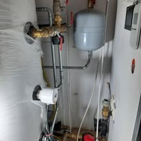 Szczegółowe ujęcie instalacji hydraulicznej z szarym zbiornikiem wyrównawczym, mosiężnymi zaworami i białym termostatem w małym pomieszczeniu technicznym.