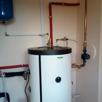 Biały, cylindryczny bojler z czarnym blatem i licznymi rurami miedzianymi oraz czerwonymi, umieszczony w rogu pomieszczenia o jasnych ścianach.