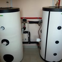 Dwa białe, cylindryczne zbiorniki z czarnymi pokrywami, połączone rurami. Widoczne elementy instalacji hydraulicznej i manometry. Ustawione przy beżowej ścianie.