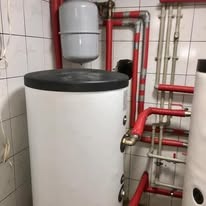 Biały, cylindryczny zbiornik buforowy z czarną pokrywą i czerwonymi rurami w tle na tle białych płytek w łazience. Elementy instalacji CO.