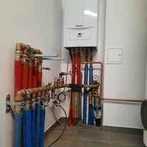 Biały piec gazowy wiszący na ścianie, z licznymi rurami miedzianymi i plastikowymi w kolorach czerwonym i niebieskim, tworzącymi skomplikowany system instalacji.