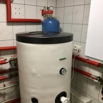 Biały bojler z czarną pokrywą i niebieskim zbiornikiem wyrównawczym w kotłowni z czerwonymi rurami na tle białych płytek. Widoczne zawory i manometry.