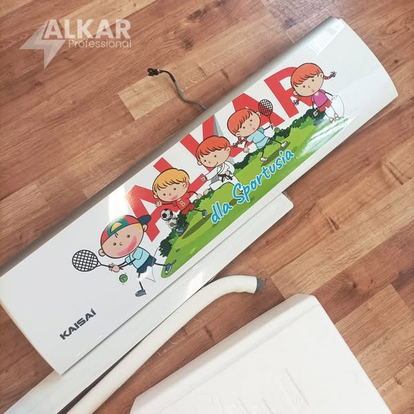 Panel klimatyzacji Kaisai z motywem sportowym dla dzieci, leżący na podłodze. Widoczny kabel zasilający i elementy montażowe. Grafika ALKAR Professional w tle.