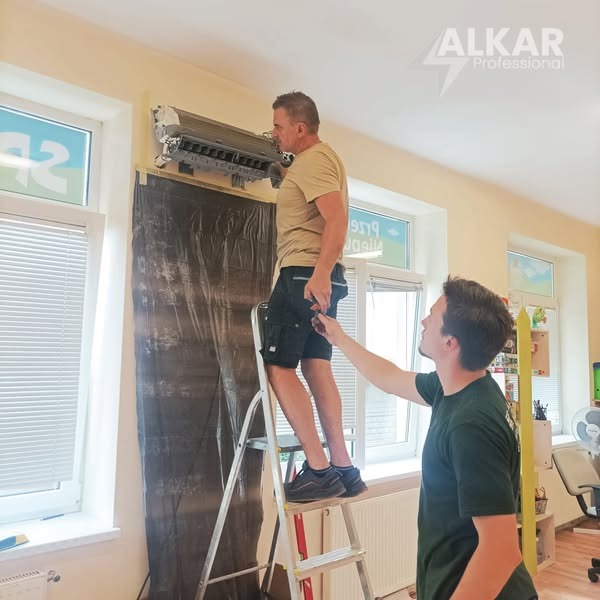 Dwóch monterów instaluje klimatyzator ścienny w pomieszczeniu z oknami, używając drabiny. Jeden z nich trzyma przewód. Widoczne logo firmy Alkar Professional.