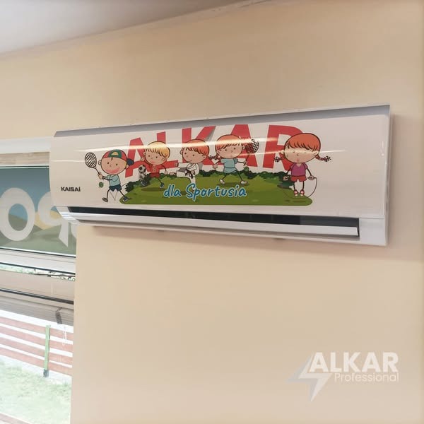 Klimatyzator Kaisai z grafiką dzieci uprawiających sport, napis 'dla Sportusia', zamontowany na ścianie. Widoczne logo ALKAR Professional.