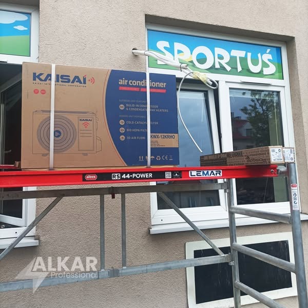 Pudełko klimatyzatora Kaisai na rusztowaniu przed budynkiem z napisem 'SPORTUS' w oknie, sugerujące montaż klimatyzacji w obiekcie sportowym.