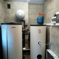 Dwa białe zbiorniki buforowe w kotłowni z szarymi płytkami, widoczne rury i elementy instalacji, mały niebieski zbiornik wyrównawczy na ścianie.