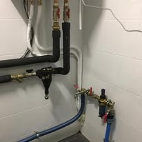 Złożona instalacja hydrauliczna z rurami, zaworami i filtrami w pomieszczeniu z kafelkami, widoczne połączenia i izolacje, niebieskie rury na dole.