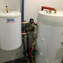 Dwa białe bojlery DIBO HOTWATER, połączone rurami miedzianymi i zaworami, na tle jasnej ściany. Instalacja hydrauliczna z widocznymi elementami przyłączeniowymi.