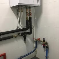 Biały piec gazowy zamontowany na ścianie z widocznymi rurami i zaworami, czarne izolacje na rurach, instalacja hydrauliczna w jasnym pomieszczeniu.