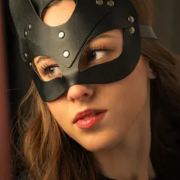 Portret kobiety w czarnej, skórzanej masce w kształcie kocich uszu, z widocznymi srebrnymi nitami, spoglądającej w dół, z delikatnym makijażem i koronkowym topem.