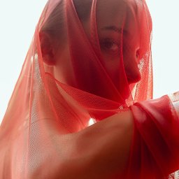 Gabriela Łasica - Portret kobiety, częściowo zasłoniętej przez czerwoną, przezroczystą tkaninę, z widocznym profilem twarzy i jednym okiem.