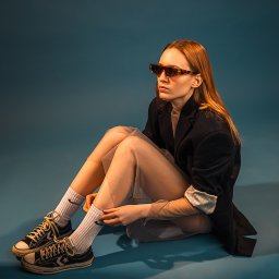 Gabriela Łasica - Kobieta siedzi na podłodze w czarnej marynarce, przezroczystej sukience, skarpetkach z logo i okularach z brązowymi oprawkami, spoglądając w bok.