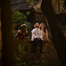 Gabriela Łasica - Chłopiec w stroju komunijnym siedzi na murku z dwiema kobietami, spoglądając w górę na tle drzew i starego budynku.