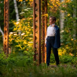 Gabriela Łasica - Chłopiec w eleganckim stroju komunijnym, z marynarką przewieszoną przez ramię, pozuje w leśnej scenerii z drewnianymi konstrukcjami w tle, oświetlony ciepłym, popołudniowym światłem.