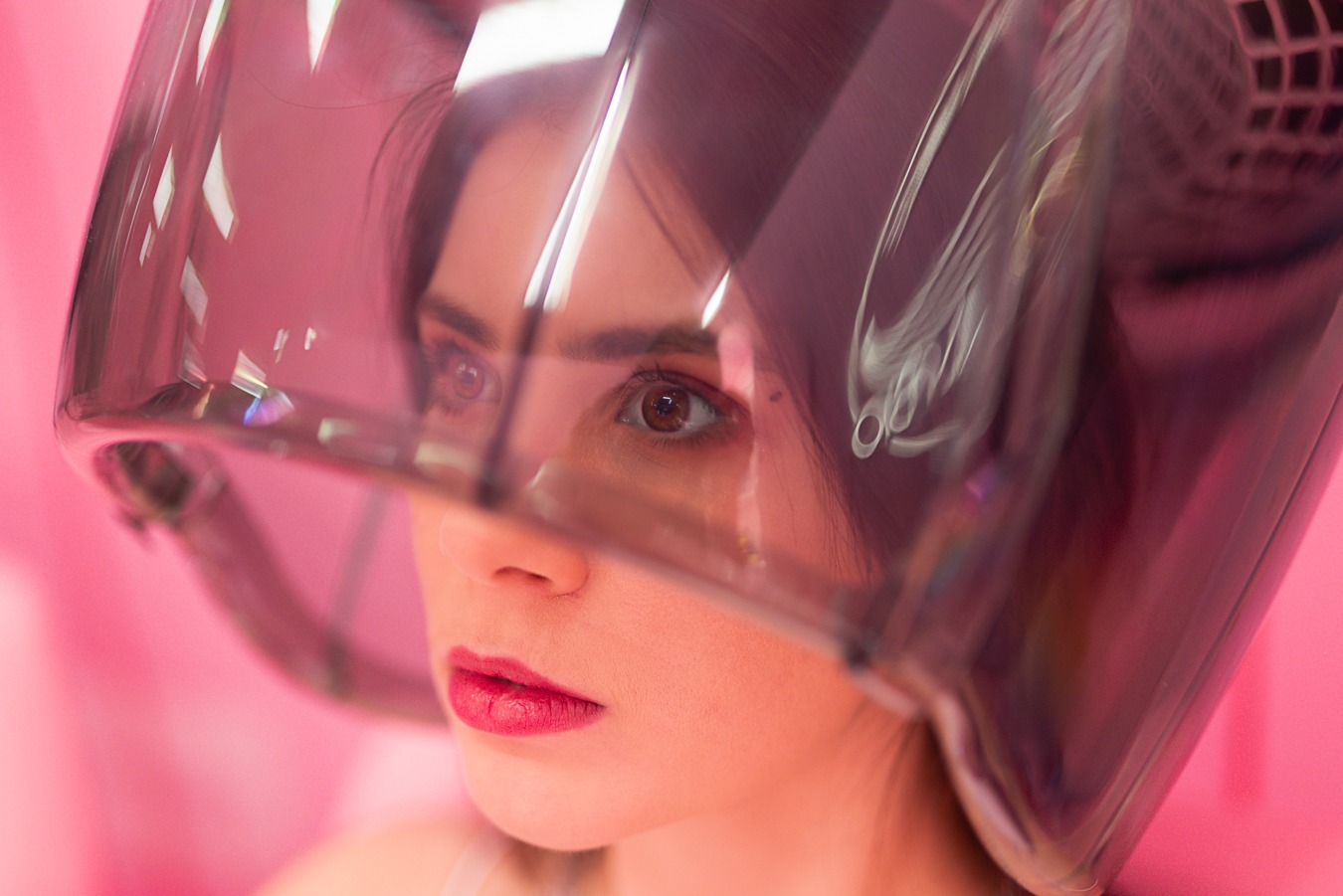Portret kobiety pod suszarką fryzjerską w salonie, z bliska widoczne oczy i usta, różowe tło.