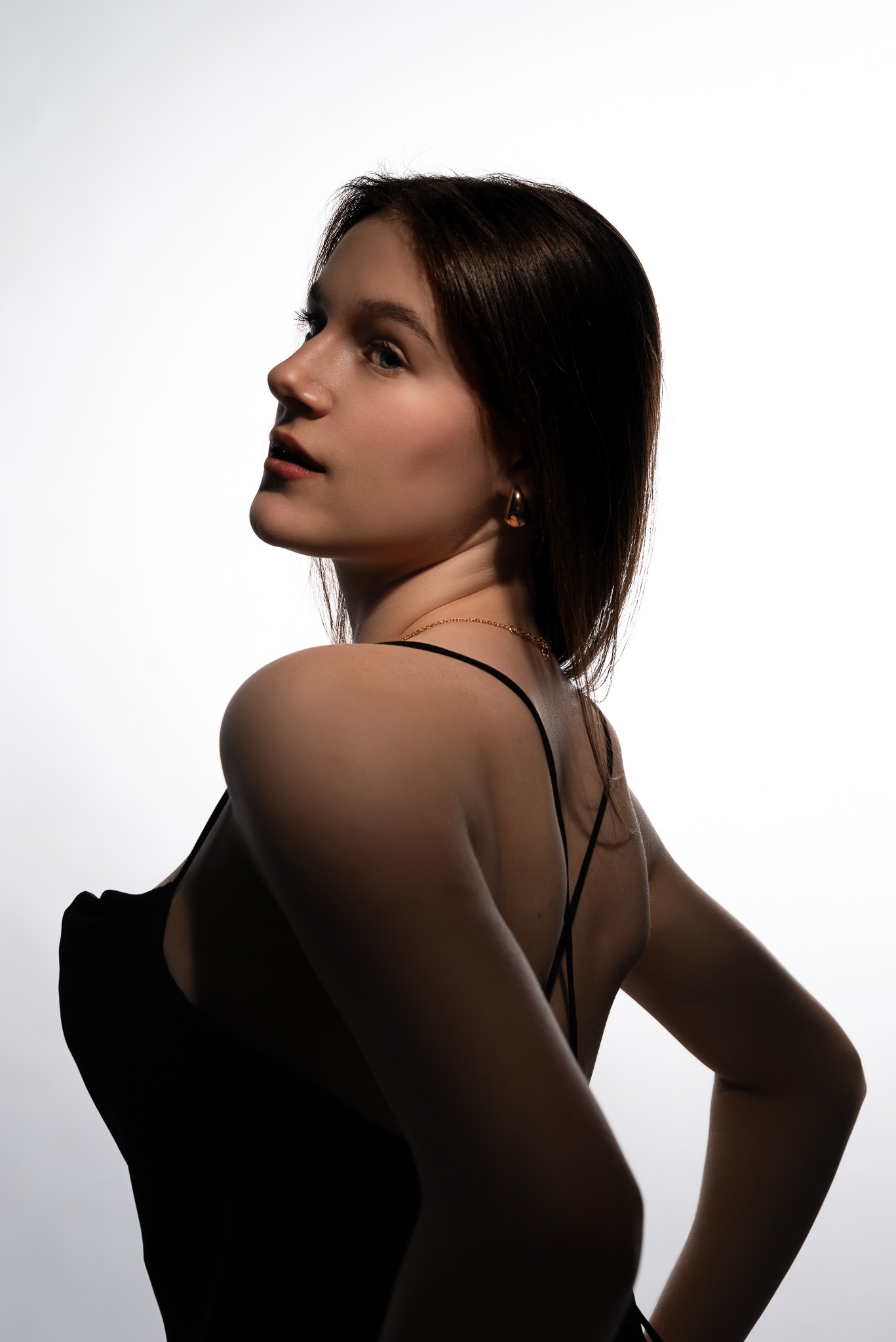 Portret młodej kobiety z profilu, z delikatnym makijażem i złotą biżuterią na tle jasnego, rozproszonego światła.