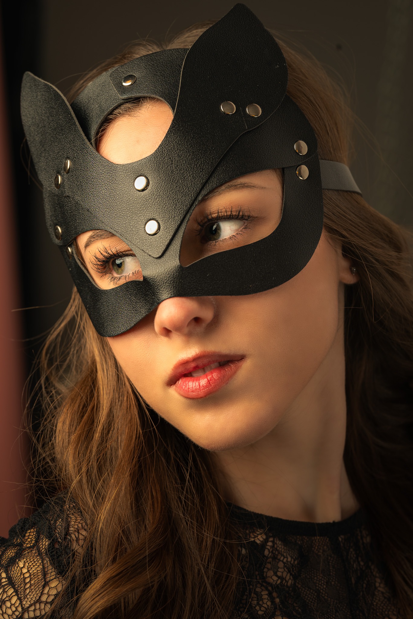 Portret kobiety w czarnej, skórzanej masce w kształcie kocich uszu, z widocznymi srebrnymi nitami, spoglądającej w dół, z delikatnym makijażem i koronkowym topem.