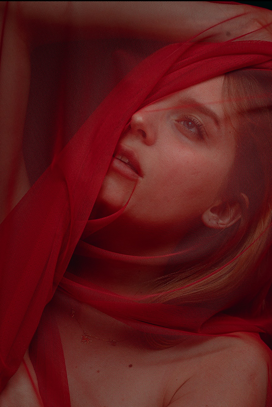 Portret młodej kobiety z delikatnym makijażem, częściowo zasłoniętej czerwoną, siatkową tkaniną, z widocznym fragmentem dekoltu i subtelnym naszyjnikiem.