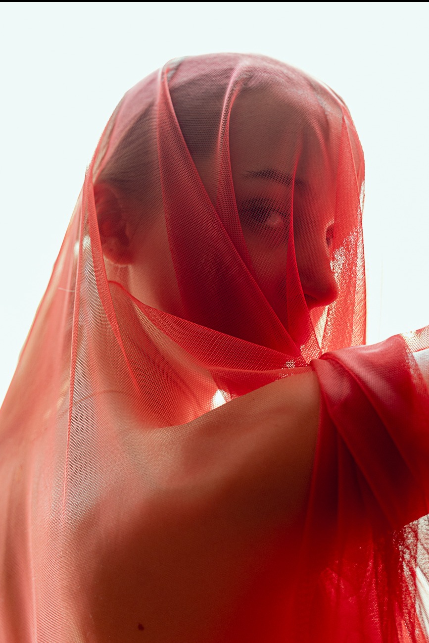 Portret kobiety, częściowo zasłoniętej przez czerwoną, przezroczystą tkaninę, z widocznym profilem twarzy i jednym okiem.