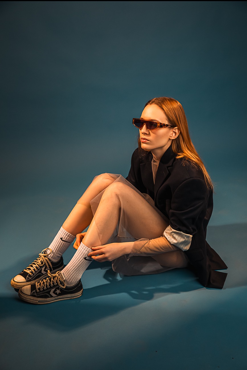 Kobieta siedzi na podłodze w czarnej marynarce, przezroczystej sukience, skarpetkach z logo i okularach z brązowymi oprawkami, spoglądając w bok.