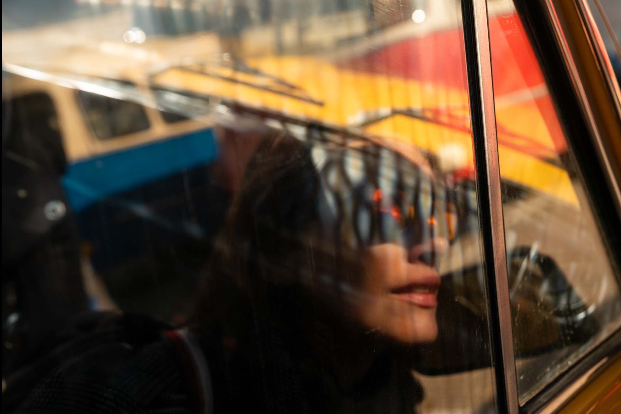 Portret kobiety odbijający się w brudnej szybie autobusu z widocznymi wycieraczkami i elementami karoserii w kolorach żółtym, czerwonym i niebieskim.