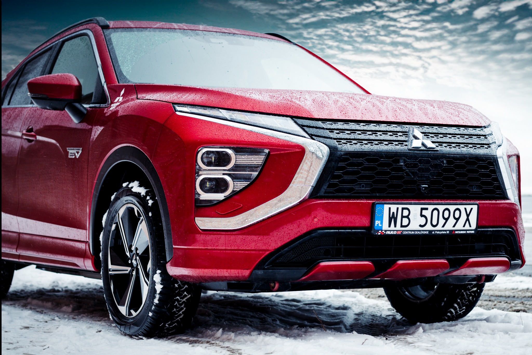 Czerwony samochód Mitsubishi Eclipse Cross z mokrą karoserią, sfotografowany z niskiej perspektywy na tle zimowego krajobrazu, z widoczną tablicą rejestracyjną WB 5099X i logo Auto GT.