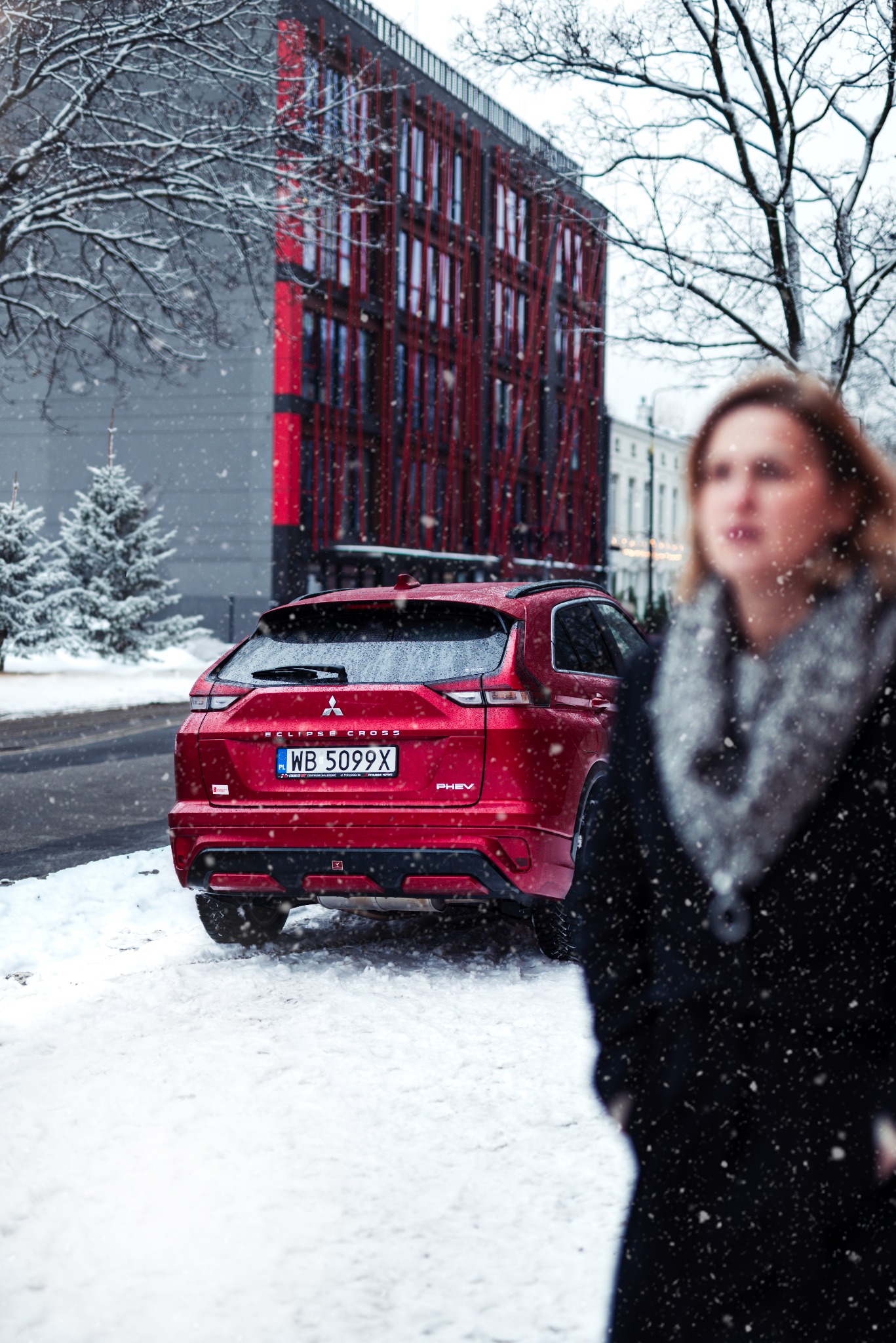 Czerwony Mitsubishi Eclipse Cross PHEV zaparkowany na śniegu w mieście, widok z tyłu, padający śnieg, budynek z czerwonymi elementami w tle, zamazana postać kobiety po prawej.