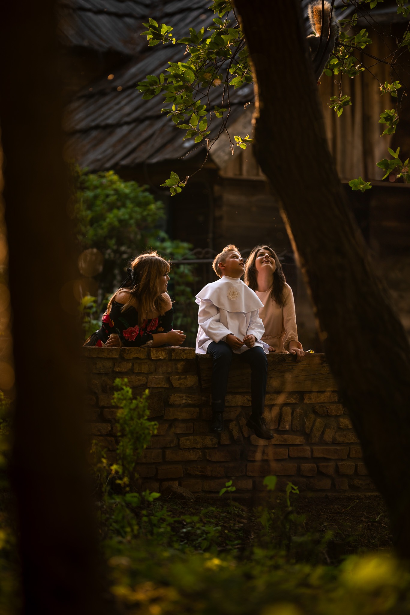 Chłopiec w stroju komunijnym siedzi na murku z dwiema kobietami, spoglądając w górę na tle drzew i starego budynku.