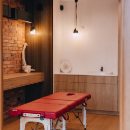 Fizjoterapia i Osteopatia Mokotów Tomasz Nowik - Czerwony stół do zabiegów w gabinecie z ceglaną ścianą, drewnianymi elementami i modelem kręgosłupa.