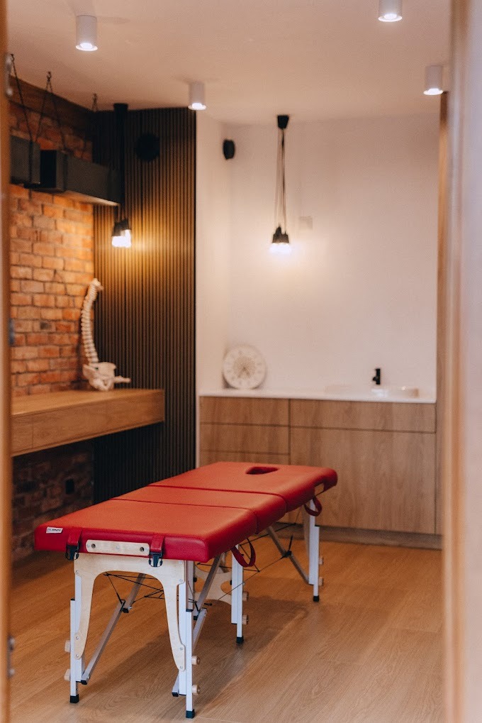 Czerwony stół do zabiegów w gabinecie z ceglaną ścianą, drewnianymi elementami i modelem kręgosłupa.