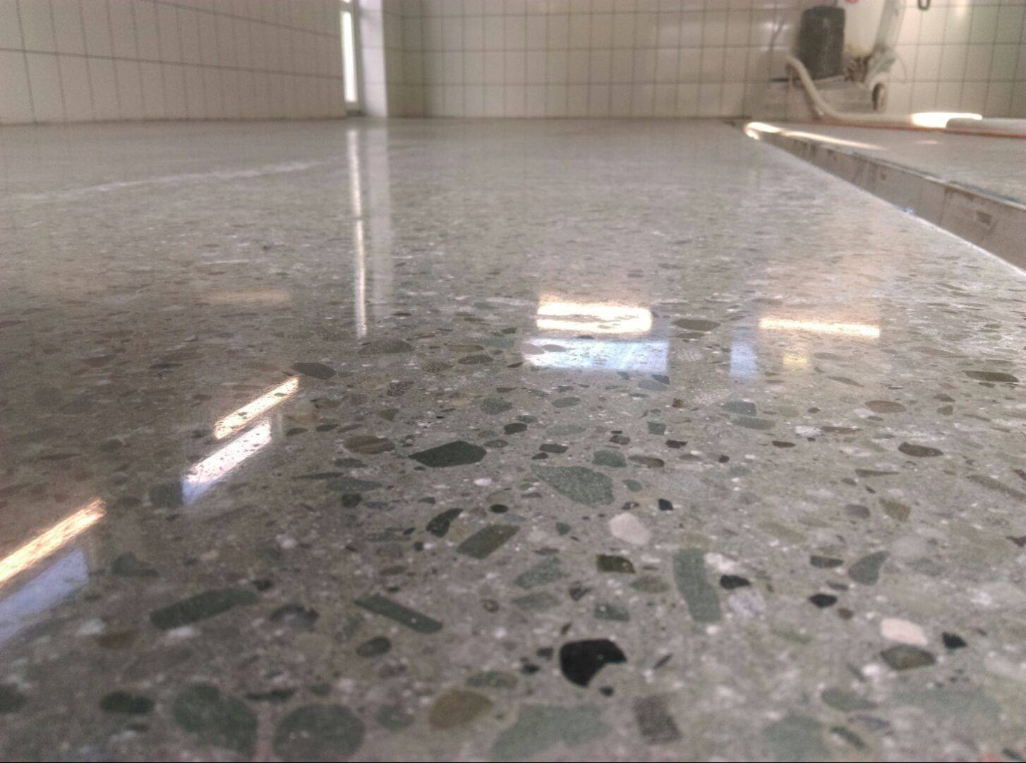 Wypolerowana, szara posadzka z widocznymi fragmentami kruszywa, odbijająca światło okna, z maszyną polerską w tle.
