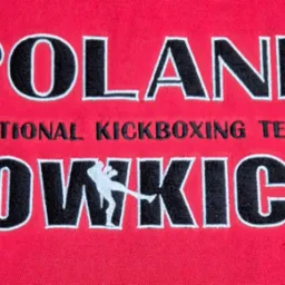 Czerwona tkanina z wyhaftowanym napisem 'POLAND NATIONAL KICKBOXING TEAM LOW KICK' i sylwetką kickboxera. Czarny tekst z białą obwódką.