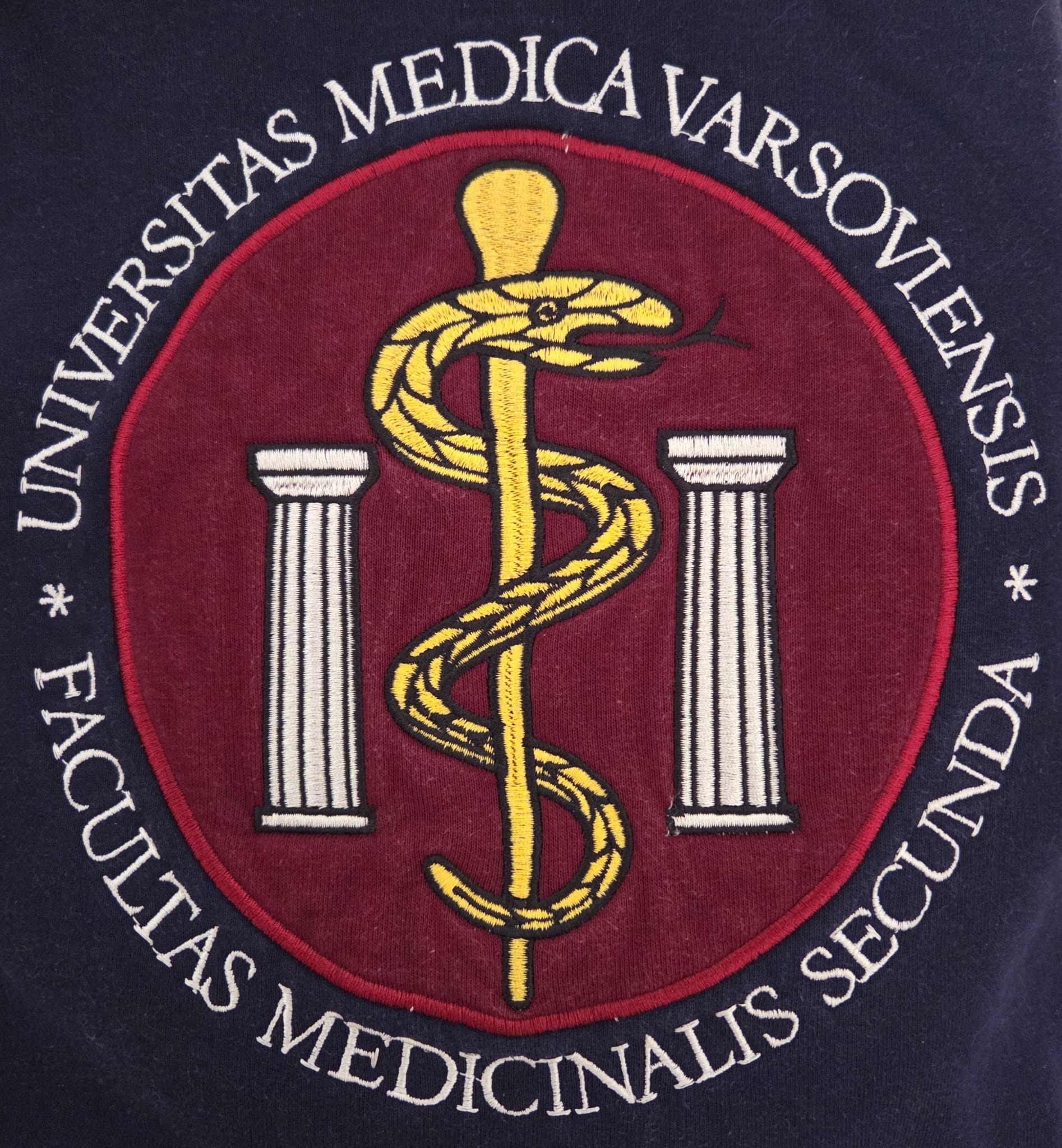 Zbliżenie na granatową bluzę z wyhaftowanym herbem Warszawskiego Uniwersytetu Medycznego, przedstawiającym laskę Eskulapa i dwie kolumny na bordowym tle.
