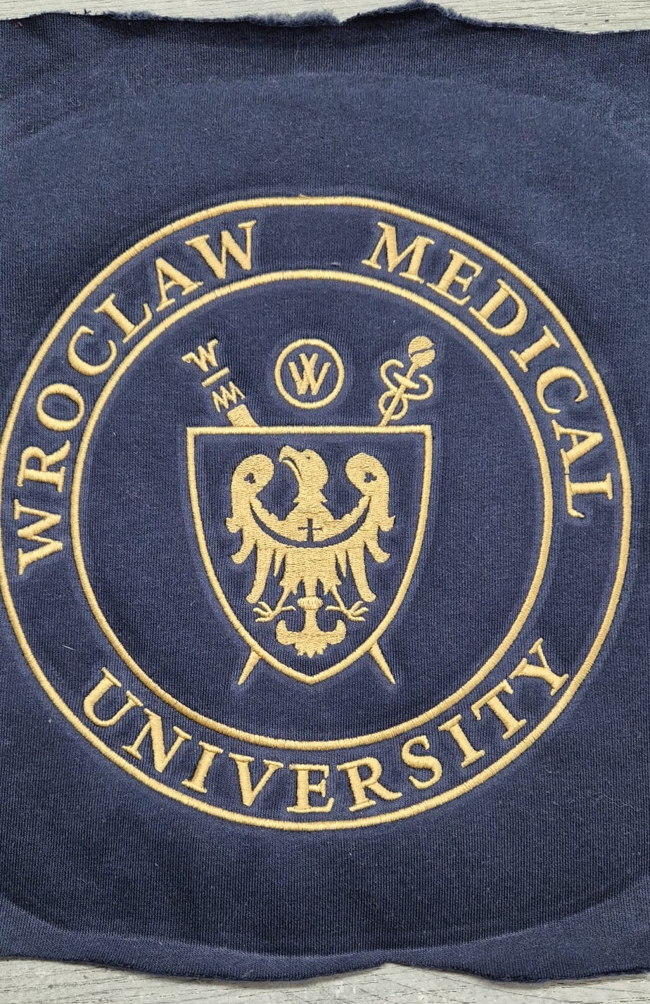 Fragment granatowej tkaniny z wyhaftowanym złotym herbem i napisem 'Wrocław Medical University' w okręgu, widoczne postrzępione krawędzie materiału.