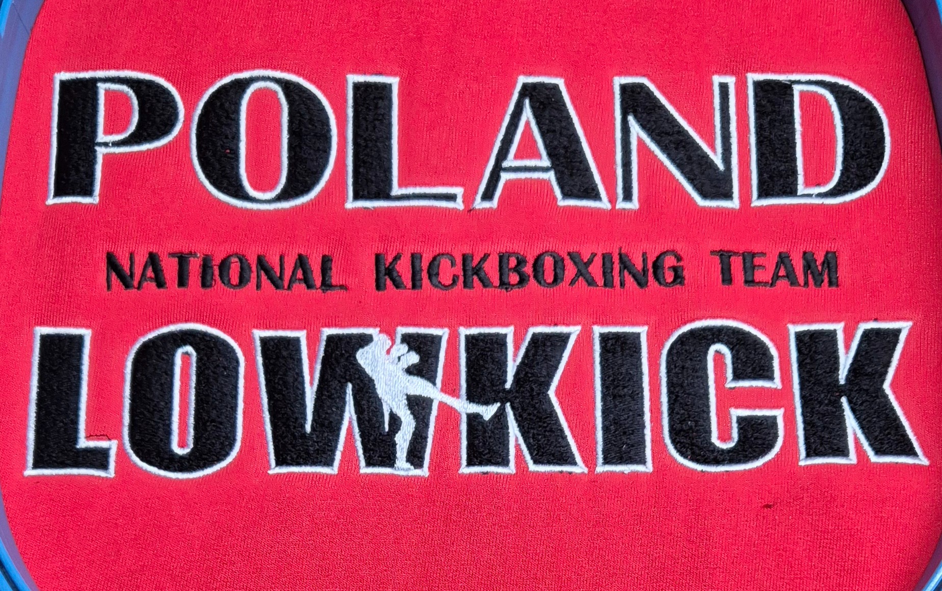 Czerwona tkanina z wyhaftowanym napisem 'POLAND NATIONAL KICKBOXING TEAM LOW KICK' i sylwetką kickboxera. Czarny tekst z białą obwódką.