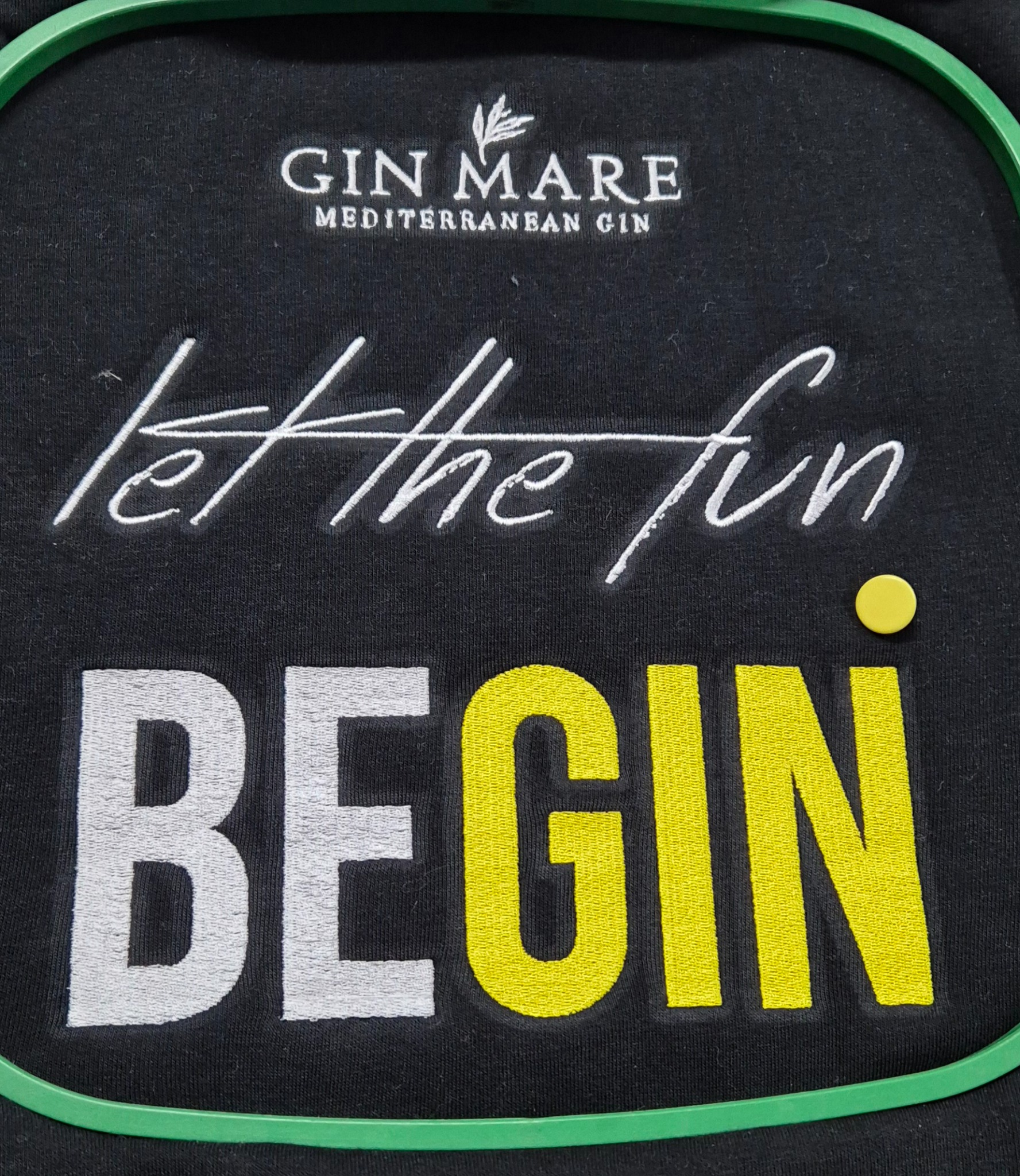 Czarna koszulka z haftowanym napisem 'GIN MARE', 'let the fun', 'BEGIN' w kolorach białym i żółtym, umieszczona w zielonej ramce do haftowania.