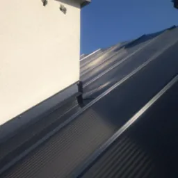 Nowoczesne, ciemne pokrycie dachowe z blachy na budynku z otynkowanym kominem, widoczne w słoneczny dzień.